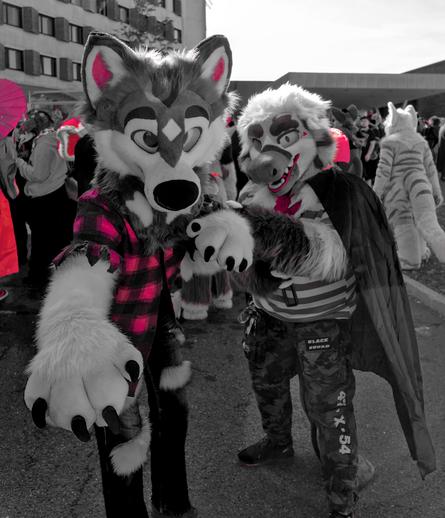 Ein Schwarzweiß Bild mit Roten Highlights.
Ein Schnabeltier in Vampir Outfit hält einen Wolf Fursuiter an einer Kette zurück