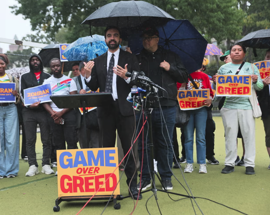 A politician wearing a dark suit with a white shirt and plaid tie stands under an umbrella with a person behind him next to a sign that says GAME OVER GREED and then there is a row of people behind him holding does signs and other signs that say ZOHRAN.