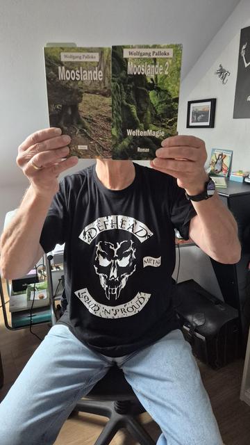Das Bild zeigt den Autor Wolfgang Palloks von vorne, der auf einem Stuhl sitzt und sich seine beiden bereits veröffentlichten  Mooslande-Romane vor den Kopf hält.