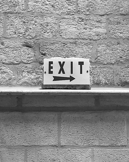A simple black and white photograph shows a sign with 'EXIT' and an arrow, attached to a stone and resting on a ledge in front of a brick wall.