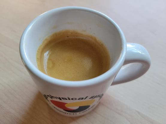 A white, 3 ounce, ceramic espresso (demitasse) cup with a shot of espresso, on a maple desktop. The blue text on the cup reads "Cyber-Physical Systems Research Center" and wraps in a circle around the logo, which looks like a loosely interlaced, rounded triquetra with thick knots in solid red, yellow, and blue (each its own color). 