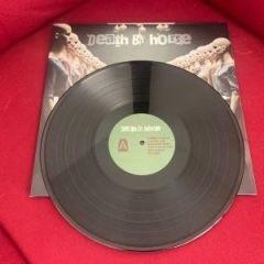 Eine Schallplatte mit roter Oberfläche und grünem Etikett liegt auf dem Albumcover mit der Aufschrift "Death & House". Auf dem Albumcover sind Bilder zu sehen, die mit dem Titel in Verbindung stehen.
