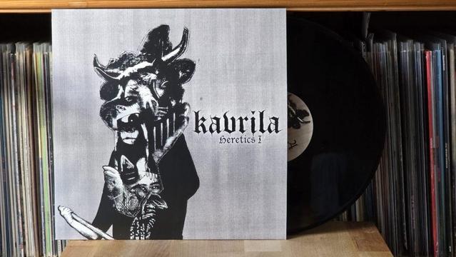 Eine Vinyl-Schallplatte mit einem Albumcover mit dem Titel "Kavrila - Heretics I". Das Cover zeigt eine künstlerische, monochrome Illustration einer Figur mit einem tierähnlichen Gesicht und stilisierten Händen. Die Schallplatte befindet sich zwischen anderen Vinyl-Alben.