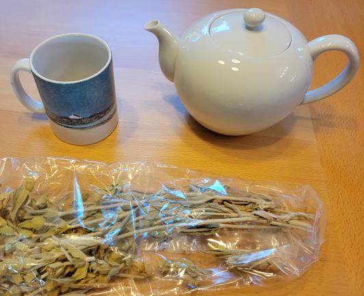 A packet of dried mountain tea herbs pictured with a mug and white tea-pot