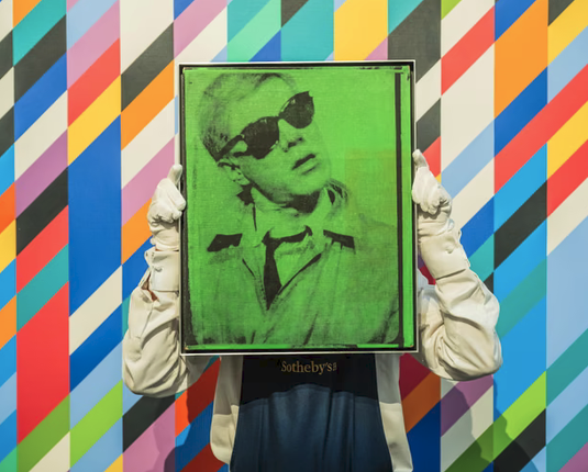 A person who holds up a painting or othr image of another person in black and green who's wearing a coat with a big collar and has dark sunglasses and his head cocked to the side.  Behind it there are a bunch of trapezoidal brightly color shapes stacked together