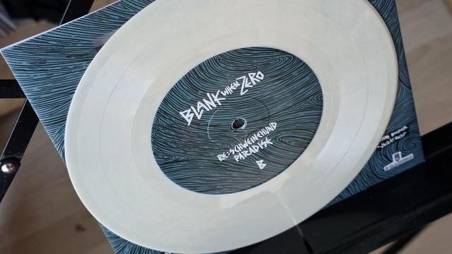 Eine durchsichtige Vinyl-Schallplatte wird mit einem Cover mit blauen Wellenmustern präsentiert. Die Platte ist auf einer Seite mit "BLANK when ZERO" beschriftet.