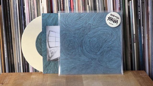 Ein Albumcover mit einem gewirbelten blauen Design wird neben einer Schallplatte und einem Schwarz-Weiß-Foto eines Fensters gezeigt. Im Hintergrund ist eine Sammlung von Schallplatten zu sehen.