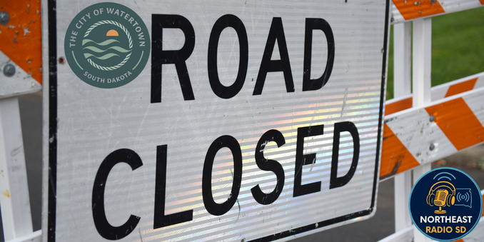 Road closure sign with bold text "ROAD CLOSED." Orange and white stripes and logos for Watertown and Northeast Radio SD visible.
