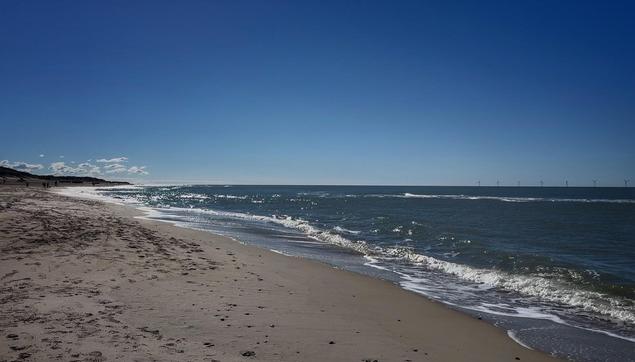 Bild eines sonnigen Nordseestrandes mit wenig Brandung. Das dunkelblau schimmernde Meer glitzert und reflektiert in Teilen die Sonne. 