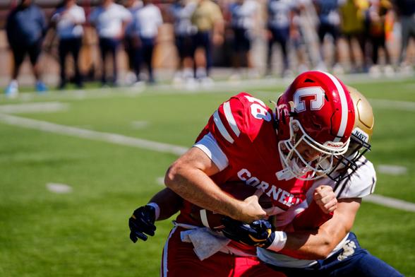 In the foreground a football player all in red with white lettering C and Cornell, 18 and stripes is carrying the ball and charging from left to right and is being grabbed from behind by a football player who you can barely see because he's behind the first player but you can see a bit of gold helmet and white jersey and blue pants,  behind a green field subdivided by a white stripe running diagnoally and a number of people with dark pants and white shirts can be seen on the far sideline out of…