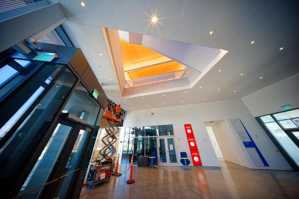 We're on the ground floor of a building with a wood floor,  to the left is a large-looking black framed door with a man riding a scissors lift to do finish work, a glass-fronted room and a row of video signs are on the far wall as well as a hallway leading back,  to the right is an exit,  above there's a hole in the ceiling showing a triangle of yellow lit ceiling bounded by a stairway spiralling around,  the near ceiling has numerous bright lights,  the closest one of which has a glorious 10-p…