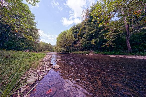 Wide angle shot of a brown looking creek with a rocks visible under the ripples,  to the left the bank has a thine row of rocks backed by some short grassy vegetation and then some deciduous trees,  that far side looks similar,  the sky is mostly blue with some blown-out white clouds.