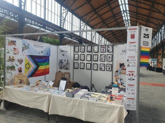 Photo de notre stand. On y voit deux tables allignées dans la longueur recouvertes d'une nappe claire. Sur laquelle sont disposés l'ensemble de nos livres. Sur la droite perpendiculaire au deux autres, une table recouverte d'une nappe foncée. Elle sera notre espace pour dédicacer nos livres.

Derrière le stand une exposition de notre livre "vent de face" et de notre livre "kléa apprend le consentement".

Sur la cloison de gauche est affichée la banderole de nos editions et dessous, un drapeau LGBT.
