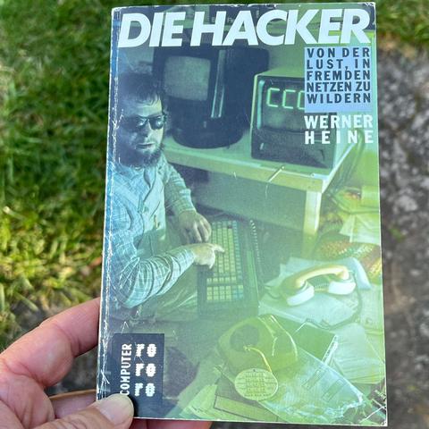 Titelseite des Buches "Die Hacker: Von der Lust, in fremden Netzen zu wildern" von Werner Heine, erschienen 1985 . Das Bild zeigt einen jungen Mann mit Sonnenbrille und Vollbart, Blick in die Kamera, Zeigefinger auf einer Tastatur. Im Hintergrund zwei Bildschirme, auf einem steht in großen grünen Buchstaben "CCC". Im Vordergrund ein Wählscheibentelefon mit abmontierter Wählscheibe. Daneben ein Telefonhörer, der in den Muffen eines Akustikkopplers steckt. Das ganze Bild ist von einem transparent…