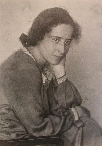 Portrait of Hannah Arendt in 1924.

Unknown author - Young-Bruehl, Elisabeth Yale University Press Hannah Arendt: For Love of the World ISBN: 978-0-300-10588-9.

Hannah is facing slightly toward the camera, but her gaze is a bit off to one side—not looking straight into the lens but very near it.

Her expression is serious, thoughtful—there is a quiet intensity in her eyes. She does not appear to smile; there's a sense of introspection.

Her dress is modest, formal: a dark jacket or dress, high neckline; possibly a blouse underneath with a collar or some delicate detail but subdued.

https://en.wikipedia.org/wiki/Hannah_Arendt#/media/File:Hannah_Arendt_1924.jpg