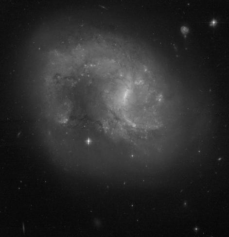 A greyscale image of face-on spiral galaxy. In the center is a small bar structure.  A single large blue spiral arm begins at the top and curves counter-clockwise from about the 11 o'clock to 9 o'clock positions. This gives the galaxy a teardrop-shape with a curved end, so that it resembles a paisley. Dark dust lanes, star clusters are scattered throughout the galaxy.