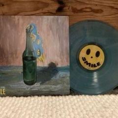 Ein Albumcover, das eine bemalte Flasche mit einem blauen Schal zeigt, die neben einer durchscheinenden blauen Schallplatte mit einem Smiley-Etikett mit der Aufschrift "SMILE" steht. Der Hintergrund ist aus Holz, und die Oberfläche ist mit einer strukturierten weißen Decke bedeckt.