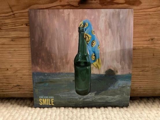 Ein künstlerisches Albumcover mit einer grünen Flasche, über die ein blaues Tuch mit Smiley-Gesichtern drapiert ist. Der Hintergrund hat eine Mischung aus warmen und kühlen Tönen, mit dem Titel "SMILE" und dem Künstlernamen "RONG KONG KOMA“.
