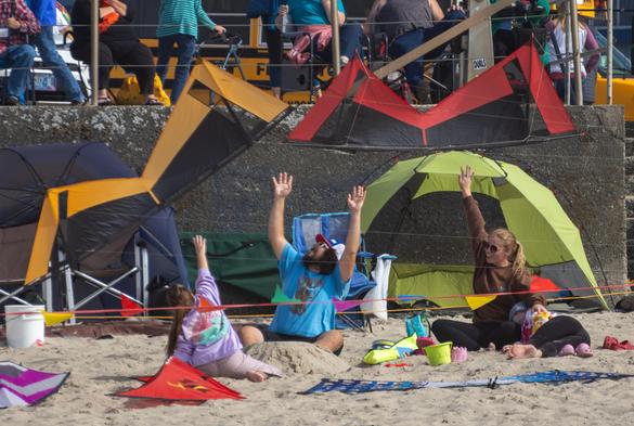 Two Essentials V quad kites doing a low inverted hover over a family on the beach. The family has their hands up in surrender, or possibly joy.
