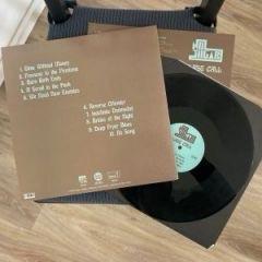 Ein Albumcover ist neben einer Schallplatte abgebildet. Das Albumcover hat einen braunen Hintergrund mit einer Liste von Titeln, und die Schallplatte ist schwarz mit einem teilweise sichtbaren Etikett. Das Bild ist auf einem Holzboden platziert.