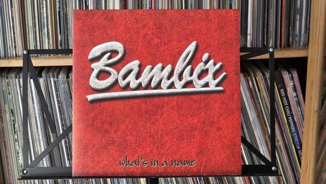 Das Bild zeigt ein Schallplattencover mit dem Titel "Bambix" in einer stilisierten Schrift auf einem strukturierten roten Hintergrund und dem Untertitel "What's in a name". Das Cover ist auf einem Ständer ausgestellt, und im Hintergrund ist eine Sammlung von Schallplatten zu sehen.