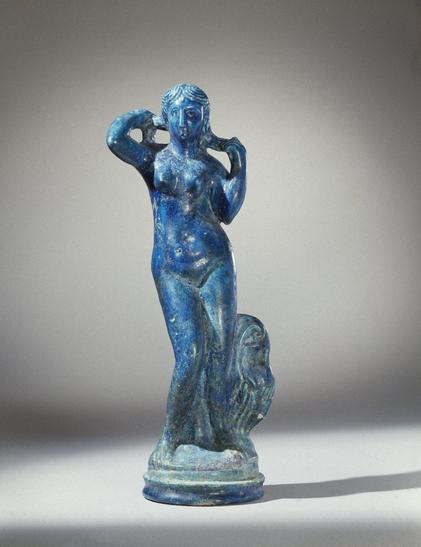 A true product of a multicultural society in Ptolemaic Egypt, this statuette combines a classical image of the Greek goddess Aphrodite appearing from the sea, just born, with the typically Egyptian material, faience. Egyptian artists achieved the characteristic brilliant blue of faience in a complex process combining ground quartz with a mixture of alkali.

The goddess is shown entirely nude, her hands raised to wring out her hair. At her left is a greatly schematized urn with garment thrown ov…