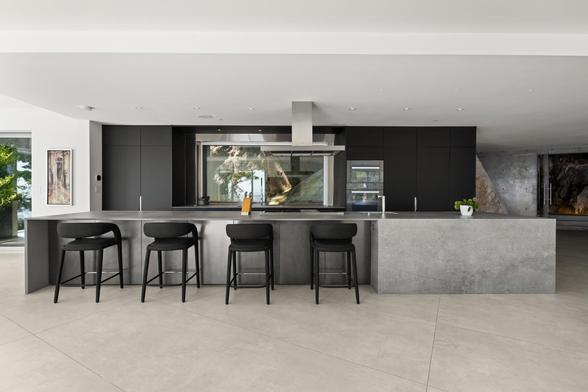 A modern kitchen in a high-end home for sale in Canada. The cabinets are dark grey, almost black. They are also flat front panel full overlay. The ginormous island is wrapped in a light grey stone, and the stools are black. The floor is off-white stone and the walls are stark white. 