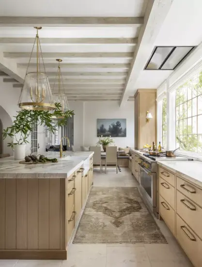 A more Contemporary, yet still leaning Traditional kitchen. The cabinets are light honey white oak with Shaker styling on the doors,but the drawers are flat front panel.  The floors are bleached wide-plank maple, and the counters are white. A traditional Persian-style runner in soft creams and browns is positioned between the oven and the island, which has two large glass and gold pendant lights above it. 