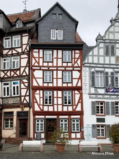 Schmales, hoch aufragendes Fachwerkhaus mit Bank und Kübel davor, eingerahmt von Nachbarhäusern.