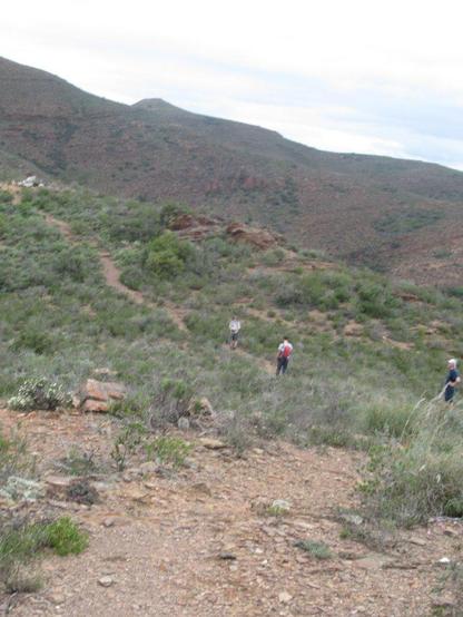 Three hikers are visible on a hiking trail through dry stoney vegetation. The path leafs toward a higher hill in the distance