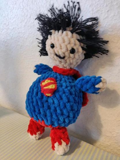 a crocheted figure in blue, red and beige that vaguely resembles a fat human being with a colored jumpsuit. The figure has an S on the chest and red cape on.