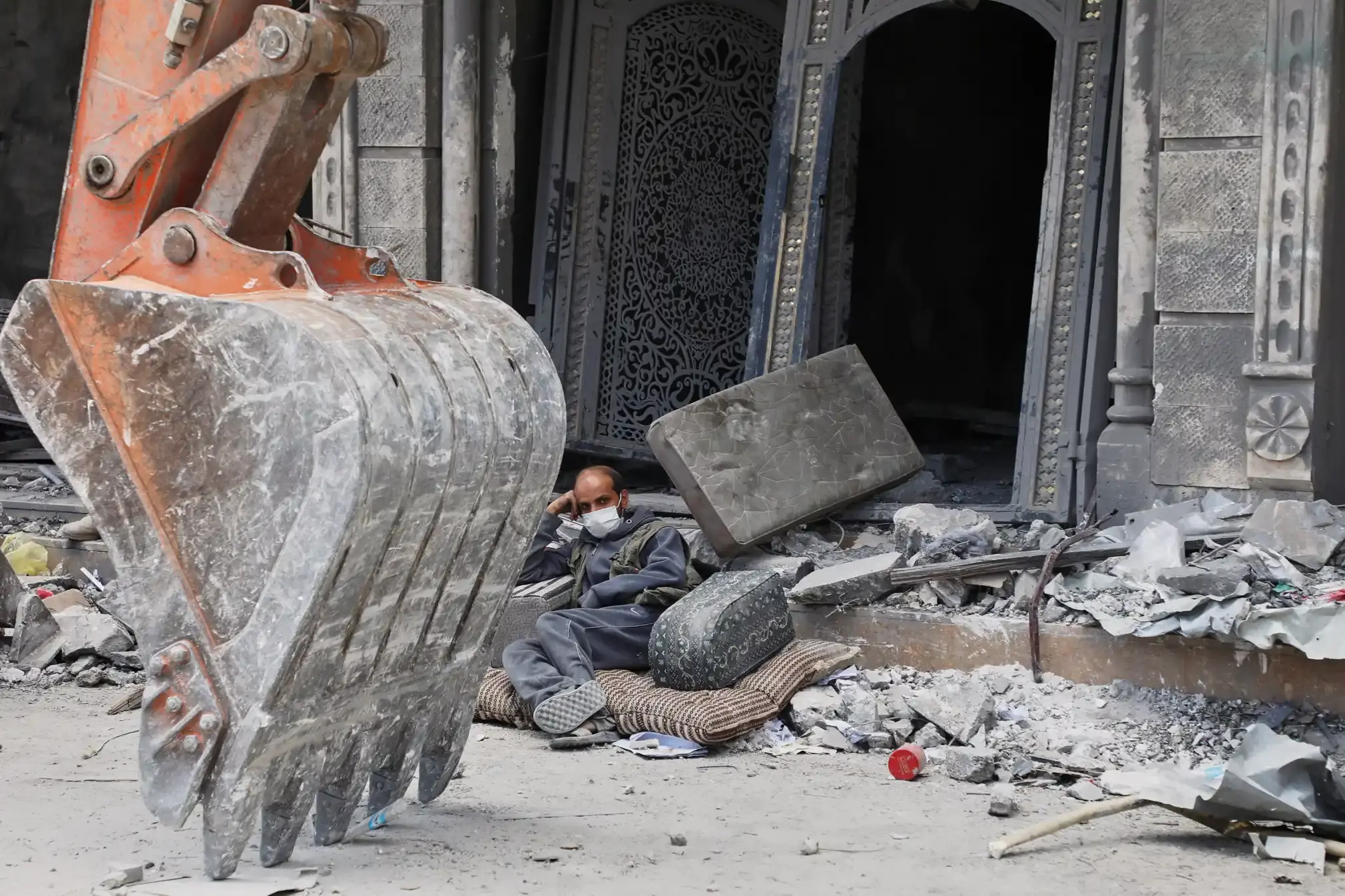 A man rests on the debris of his damaged home, next to a giant bulldozer.