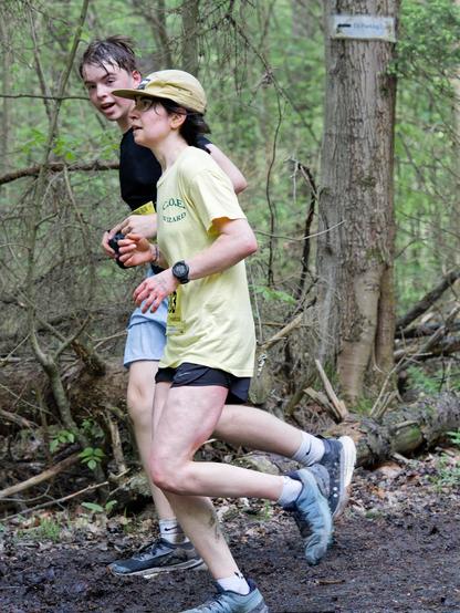 A man and woman are seen running from the side past a tree with a sign on it.  The woman is in font and has a yellow shirt and we see just a little bit of her black shorts.  Her shoes are light grey,  one shade lighter than his and she's wearing a beige billed cap and looking straight forward.  He's behind her and his making eye contact with me and has a black shirt and and blue shorts.  Both have one foot on the ground and one up and the forest is full of young spring vegetation behind them.