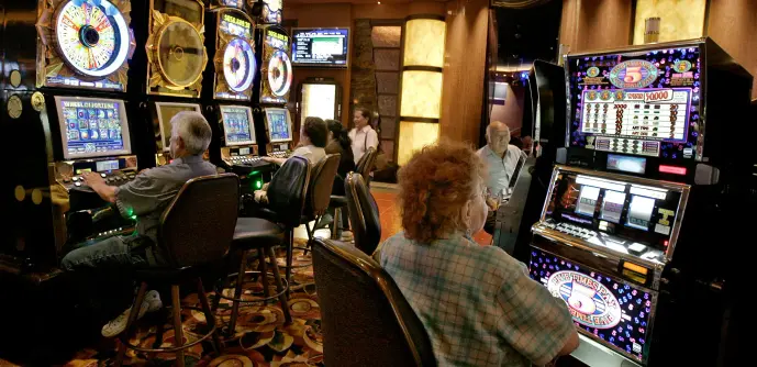 People sit and play at rows of brightly lit slot machines inside a casino.