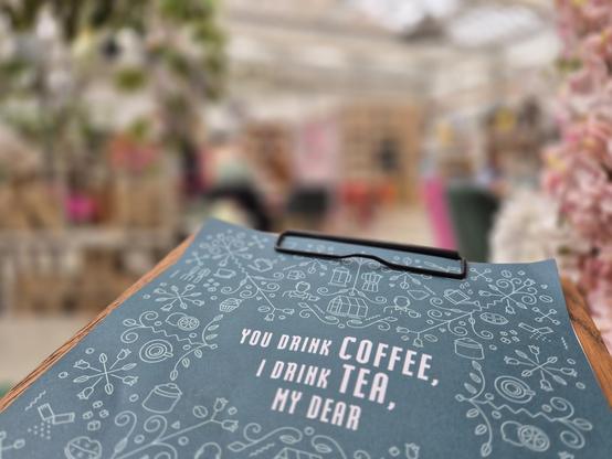 A charming, close-up photo of a clipboard menu at a cozy café or market stall, featuring a playful and welcoming message: "You drink COFFEE, I drink TEA, my dear." The menu is adorned with delicate, hand-drawn illustrations of coffee cups, teapots, and plants, adding a touch of warmth and creativity to the scene. The background is softly blurred, hinting at a lively and inviting atmosphere with greenery and colorful decorations.