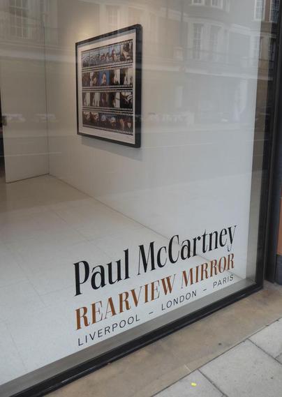 Large, plate glass window looking in on a white gallery space. On the window at the bottom are the words Paul McCartney. Rearview Mirror. London - London - Paris.