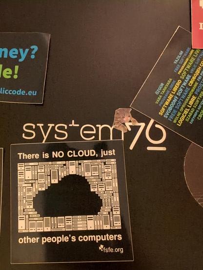 Laptoprückseite mit Stickern zu freier Software, davon zu sehen eines mit "there is no cloud, just other people's computers", das Logo (system76) des Laptops, und ein Katzensticker.