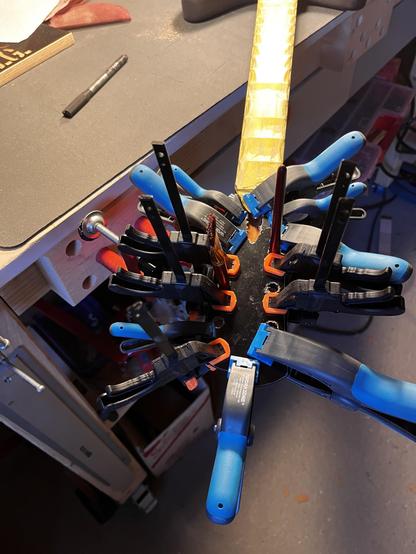 Die Kopfplatte einer E-Gitarre wird mit Hilfe von sehr vielen kleinen Zwingen aufgeleimt. Die Gitarre liegt auf einer Werkbank, der Hals ist mit gelbem Klebeband abgeklebt.