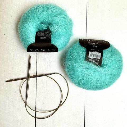 Flat lay view of 2 balls of bright mint kidsilk yarn with black labels, as well as a circular knitting needle on a white plank background.