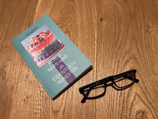 Ein Buch mit dem Titel "Klaus Nomi war ja eigentlich Konditor" von Ulli Engelbrecht liegt auf einer Holzfläche neben einer schwarzen Brille. Auf dem Buchdeckel ist eine bunte Torte abgebildet.