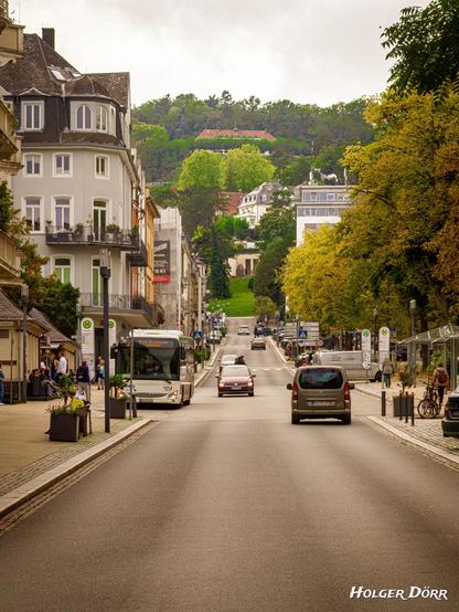 Blick entlang der Parkstraße in Bad Nauheim: Eine belebte Straße mit Bus, Autos und Passanten, gesäumt von eleganten Häusern und herbstlichen Bäumen. In der Ferne erhebt sich der Johannisberg, der Hausberg der Stadt, und verbindet den urbanen Alltag mit Natur und Geschichte.
