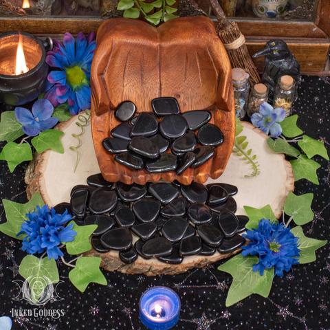 Several Apache Tears Slices rest in cupped, carved wooden hands, while other specimens spill onto a wooden surface. They are irregularly shaped and tumbled smooth in shades of black and brown. Presented by Inked Goddess Creations.