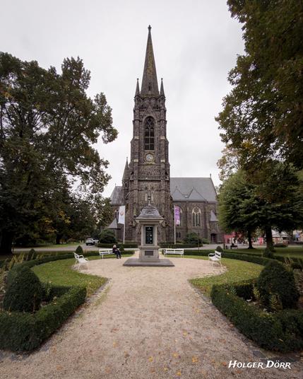 Frontale Ansicht der Dankeskirche in Bad Nauheim, einer evangelischen Kirche im neugotischen Stil aus der Zeit um 1910. Vor dem Turm führt ein Weg mit weißen Bänken und einem kleinen Schaukasten zur Kirche, flankiert von herbstlichen Bäumen.