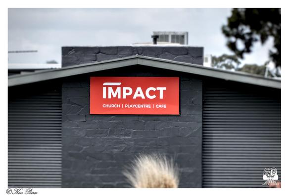 A bold red rectangular sign with the white text "IMPACT" is centered beneath the apex of a dark grey, gable-style roof. 

Below "IMPACT" in smaller text are the words "CHURCH | PLAYCENTRE | CAFE". The building's walls feature both corrugated iron and dark, textured stone.
