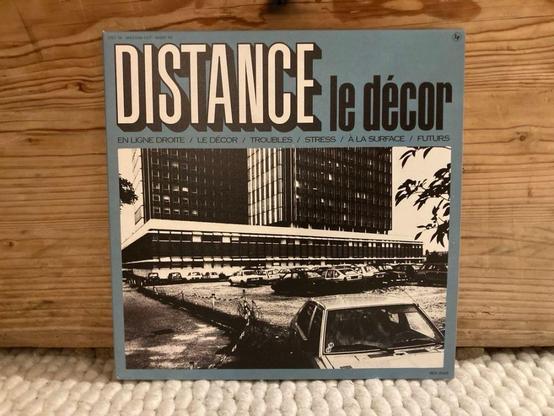 Ein Vintage-Schallplattencover mit dem Titel "DISTANCE le décor", das eine monochrome Fotografie eines Gebäudes und von Autos zeigt. Der Text enthält verschiedene Titel in französischer Sprache, die auf Themen wie gerade Linien, Dekor, Probleme, Stress, Oberfläche und Zukunft hinweisen.