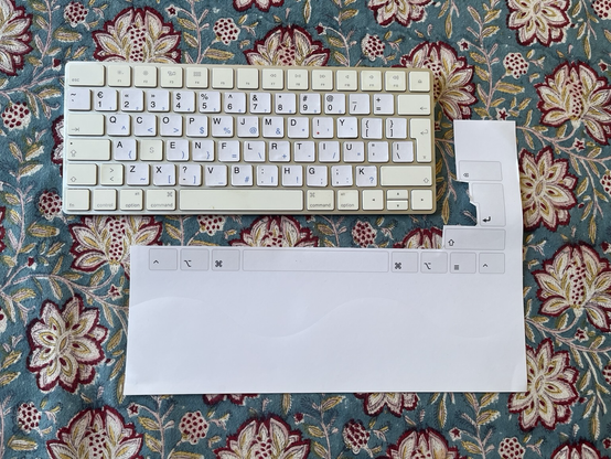 Clavier de Mac avec les touches recouvertes d’étiquettes autocollantes correspondantes à la disposition Ergo-L. À côté, le reste de la feuille autocollante aillant contenu les étiquettes. 