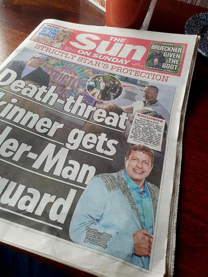 Das Bild zeigt die Titelseite der britischen Boulevardzeitung "The Sun on Sunday". Der Titel lautet "Death-threat winner gets bouncer guard" und berichtet über einen Gewinner der Show "Strictly Come Dancing", der aufgrund einer Bedrohung mit einem Todesversuch einen Bodyguard engagiert hat. Der Artikel wird von Hannah Hope geschrieben und befindet sich auf der Seite 4 und 5. Im oberen Bereich der Titelseite ist ein kleiner Artikel über "Brueckner given the boot" zu sehen. Die Zeitung liegt auf …
