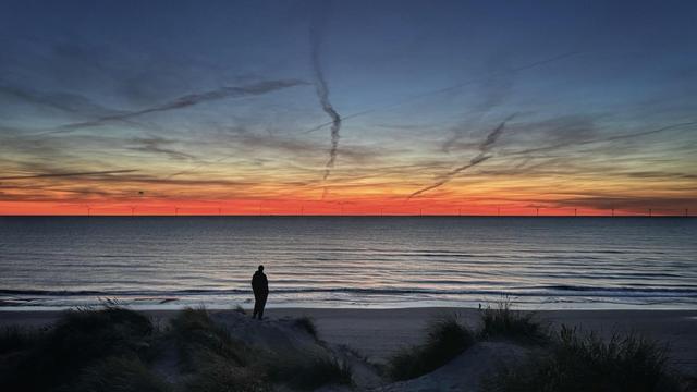 Die Silhouette einer Person in der Ferne, die auf einer hohen Düne an einem Strand steht und aufs Meer hinausschaut. Am Horizont ist die Sonne gerade untergegangen und zeigt einen orange-gelb-blauen Farbverlauf am Himmel.