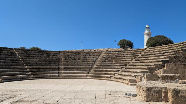 A Roman amphitheatre, devoid of people, with a white lighthouse and clumps of evergreen trees in the background to the right 