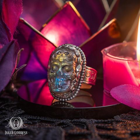 An example of the "Labradorite Etched Skull Tibetan Adjustable Ring" product sits on a black stone disc. The labradorite gemstone is a large oval carved into a skeletal face and is set in a Tibetan silver bezel. The band on the ring is adjustable to fit most fingers and engraved with swirling designs. Presented by Inked Goddess Creations.
